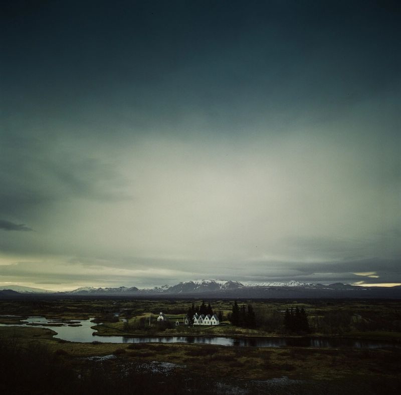 Cinematic landscape of a white chapel by a river in Iceland with distant snow-capped mountains under a dramatic sky.