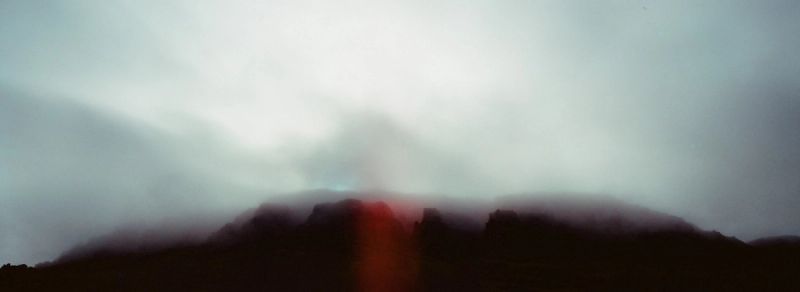 A misty mountain ridge silhouette against a cloudy sky with a red light leak, shot on Hasselblad X-Pan.