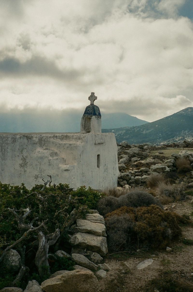 A white chapel with a cross stands on rocky terrain against distant mountains under a cloudy sky.