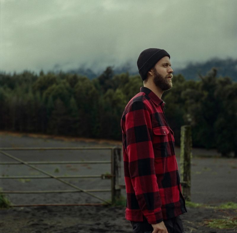 Man in red plaid shirt and beanie stands near a metal gate with a moody forest background.