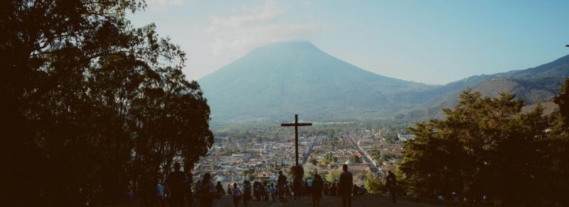 A wooden cross overlooks a town and large mountain in Guatemala, framed by trees in this film photograph.