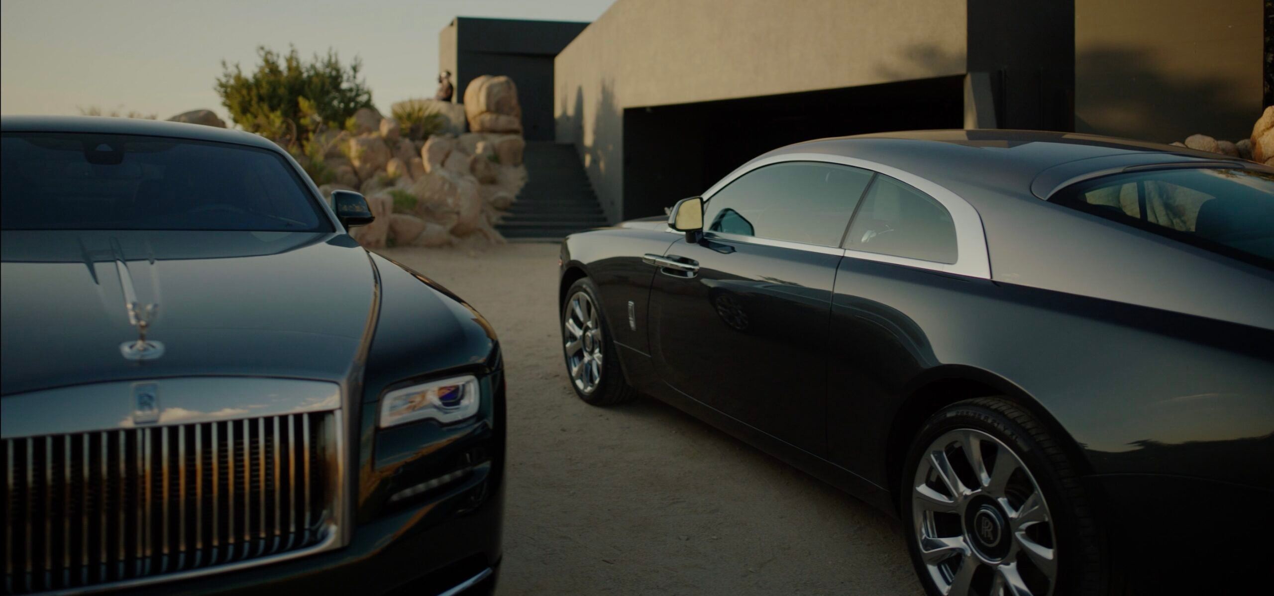 Two black Rolls-Royce luxury cars parked near a modern building with rocks in the background.