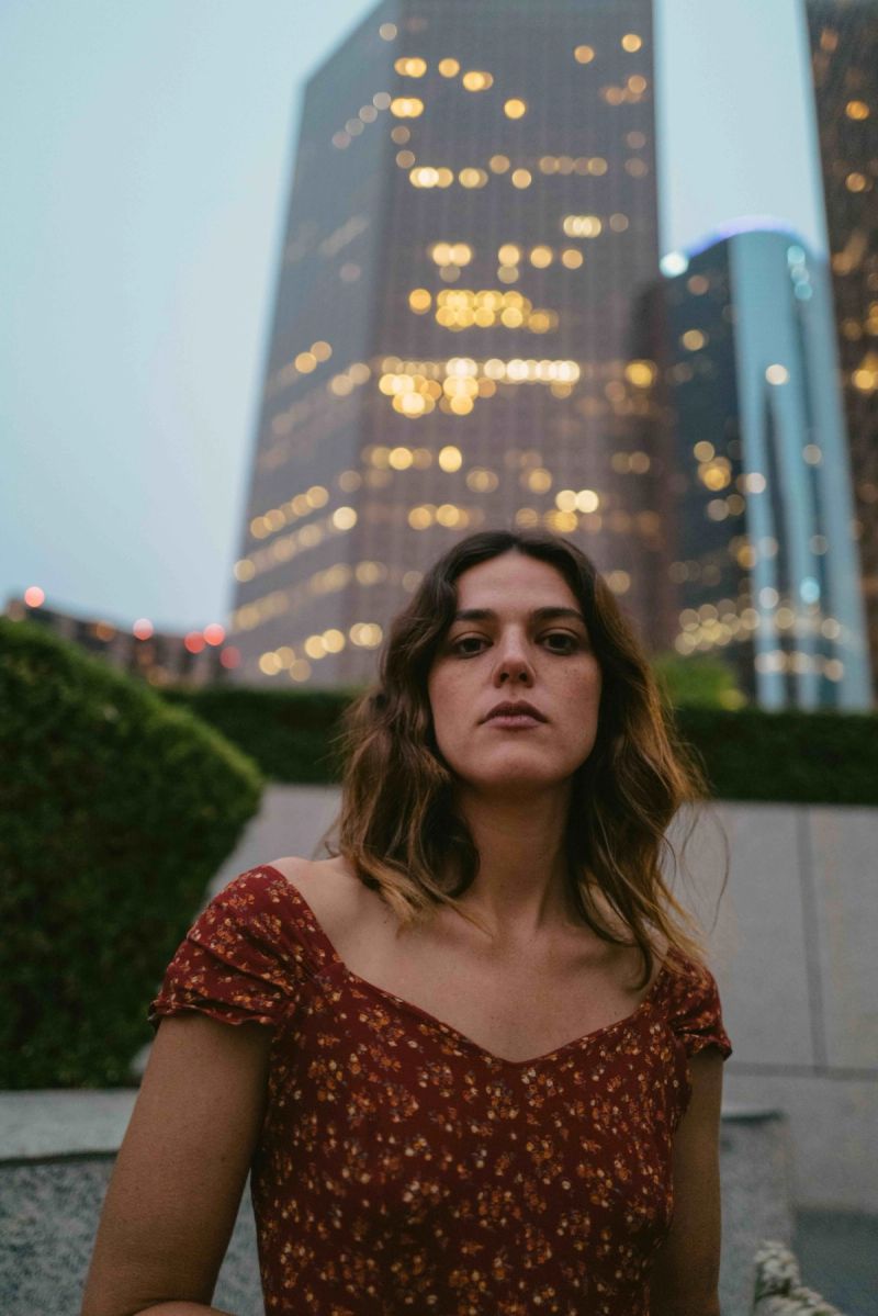 Woman in floral dress stands before city skyline at dusk