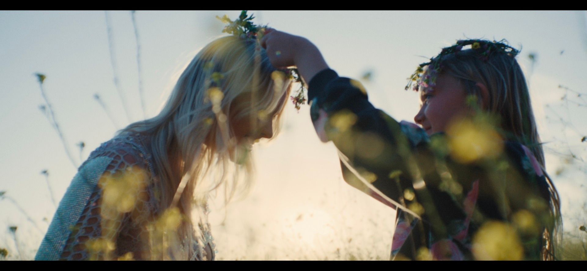 Girl placing flower crown on woman's head in sunlit field