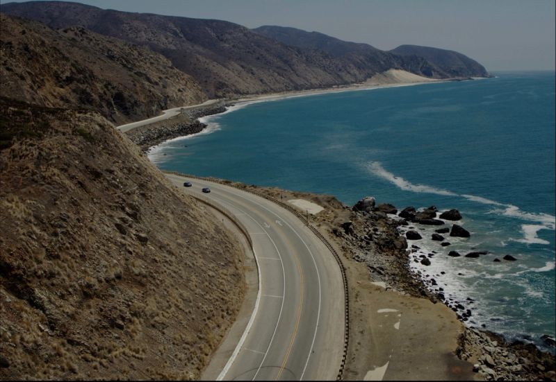 Cinematic view of a winding coastal highway with cars near the ocean.