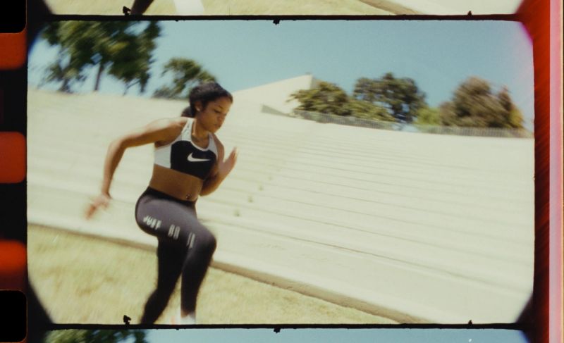 Female athlete sprinting on a track wearing Nike gear with bleachers in background.
