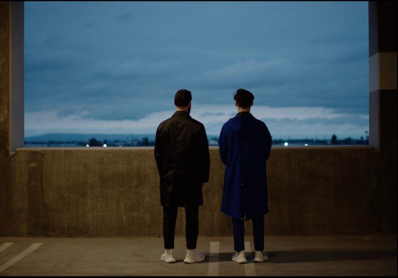 Two men in coats stand with backs to camera gazing at a twilight cityscape from an elevated concrete structure.