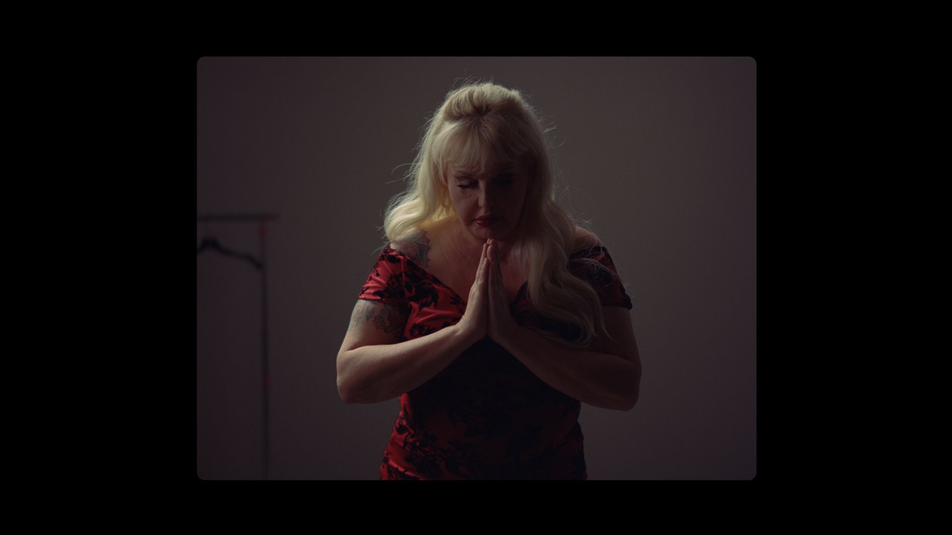 Blonde woman in red floral dress with hands clasped, dimly lit portrait
