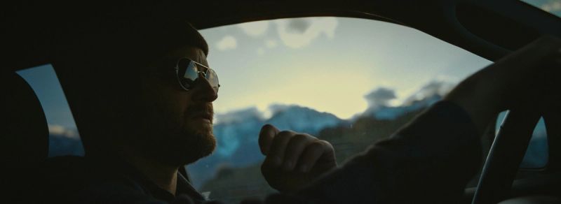 Profile of a man in sunglasses driving a car with mountains visible through the window.