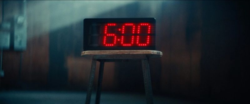 Red digital clock displaying 6:00 sits on a wooden stool in a dimly lit industrial space.