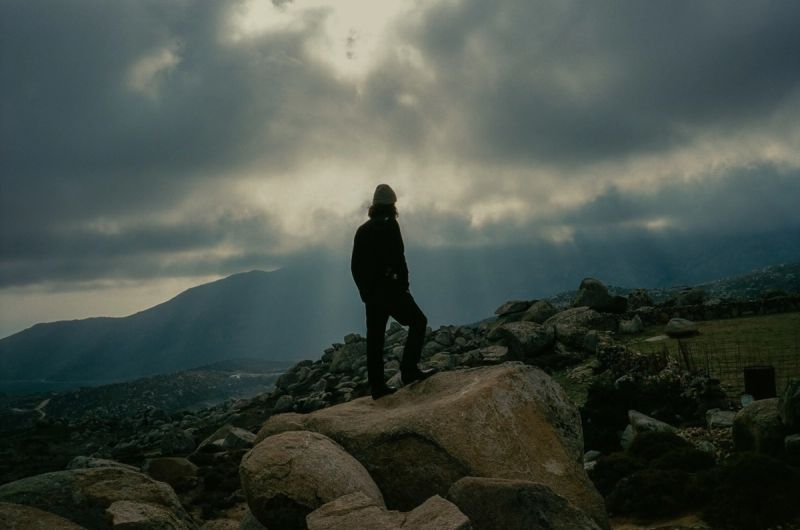 Lone figure stands on rocky peak under dramatic storm clouds in Greek islands landscape.