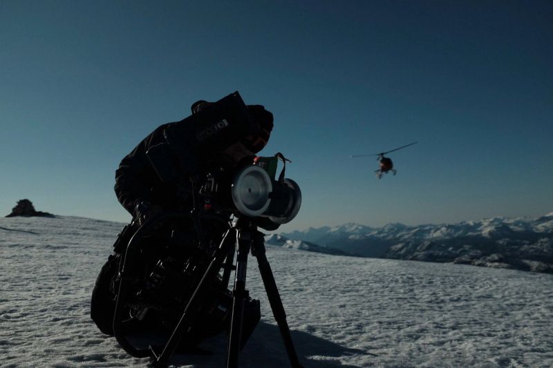 Cinematographer sets up camera on snowy mountain slope with helicopter in background