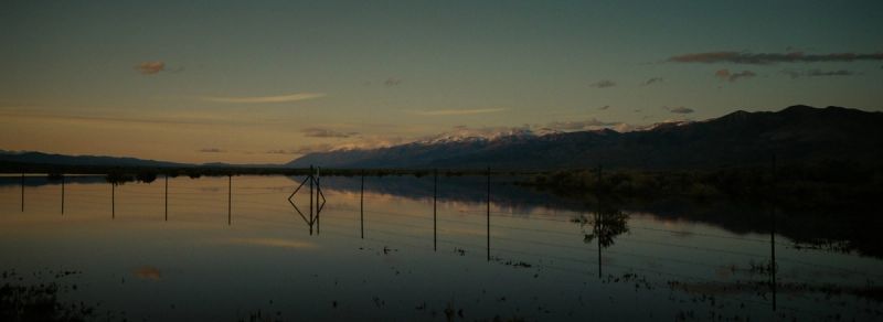 Snow-capped mountains at dusk reflected in a flooded field with a fence line.