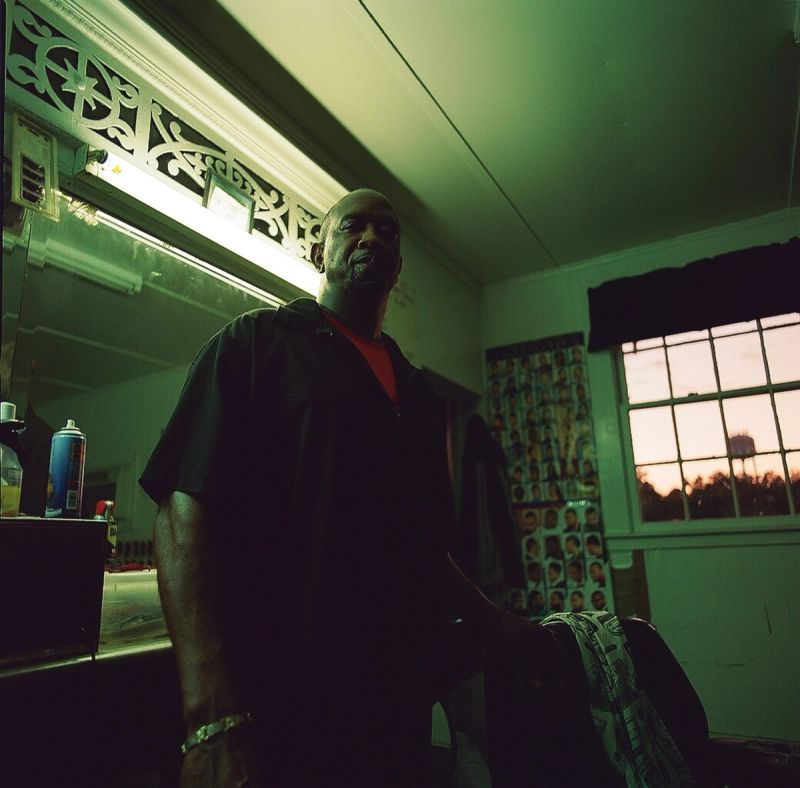 A man stands in a dimly lit barbershop with green film tones and a sunset window.