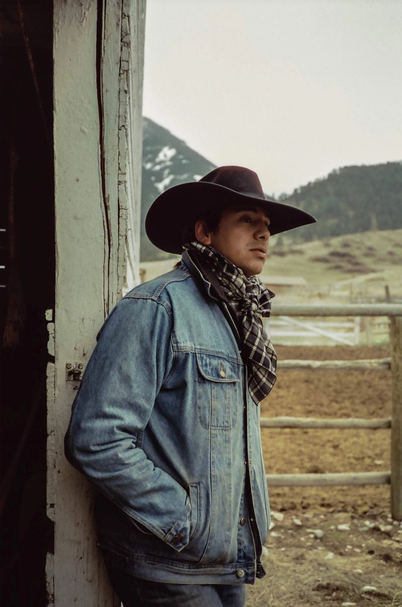 Cowboy in denim jacket and hat leans against weathered wood with mountain range background.