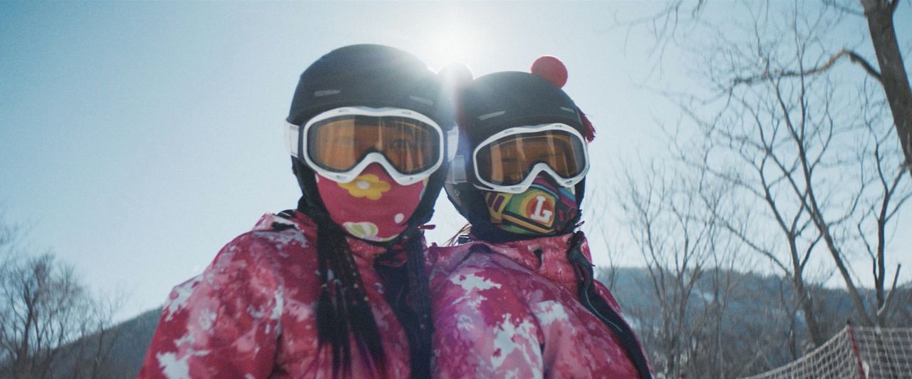 Two skiers in colorful gear and goggles pose under bright sun on snowy mountain slope.