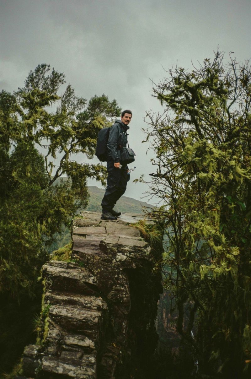 Man with backpack standing on rocky cliff edge surrounded by mossy trees in mountains.