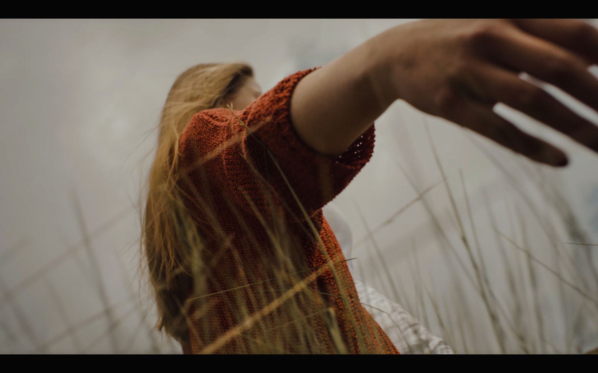 Woman in rust-colored knit sweater reaching toward sky with long blonde hair