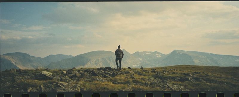 A lone figure stands on a rocky ridge overlooking the Colorado mountain range under a cloudy sky.