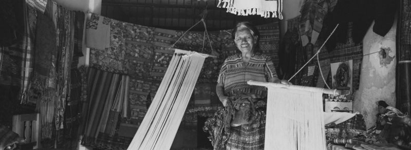 Black and white film photo of a smiling woman weaving traditional textiles on a loom in Guatemala.