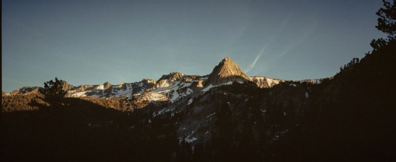 Golden hour light hits rugged mountain peaks with snow patches and pine trees.
