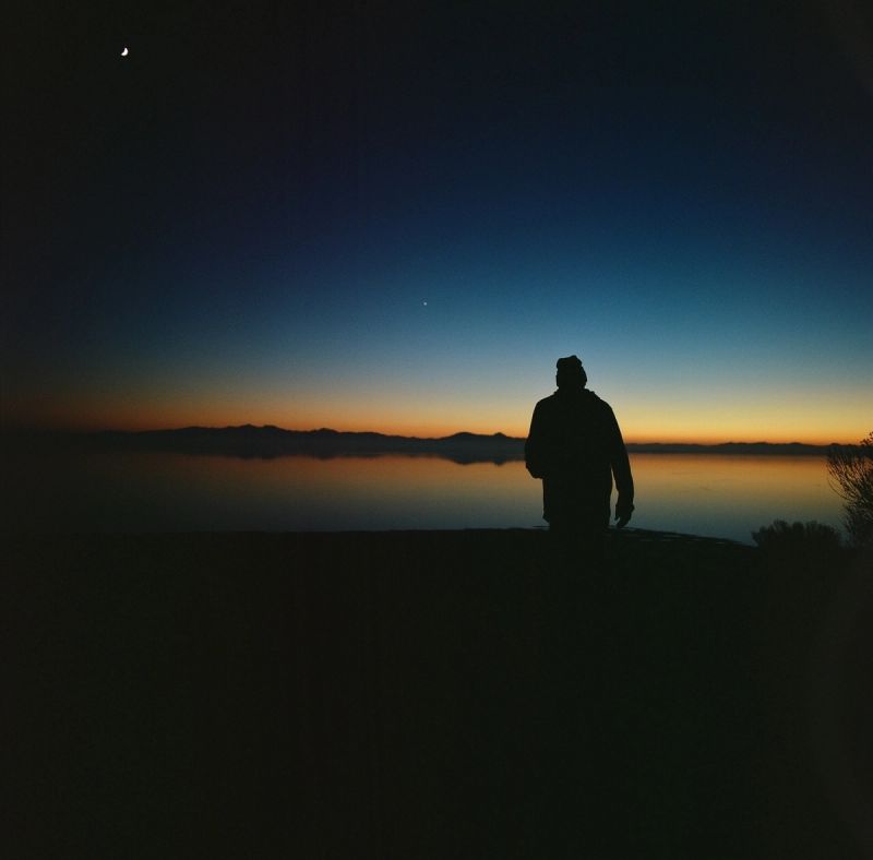 Silhouetted figure standing on a shore overlooking calm water and mountains at twilight with a crescent moon.
