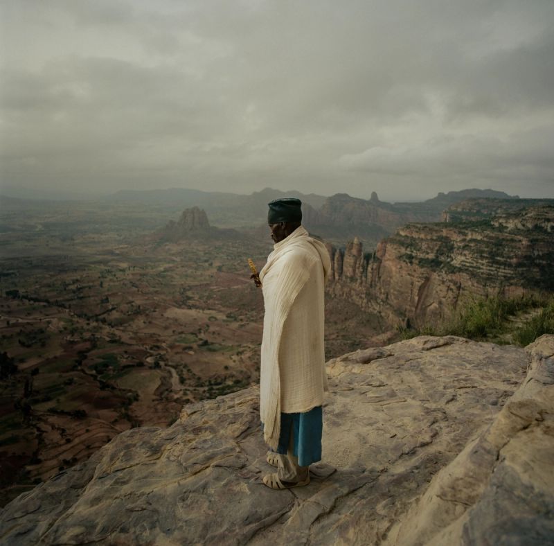 Figure in traditional attire stands on cliff edge overlooking dramatic Ethiopian valley landscape under cloudy sky.