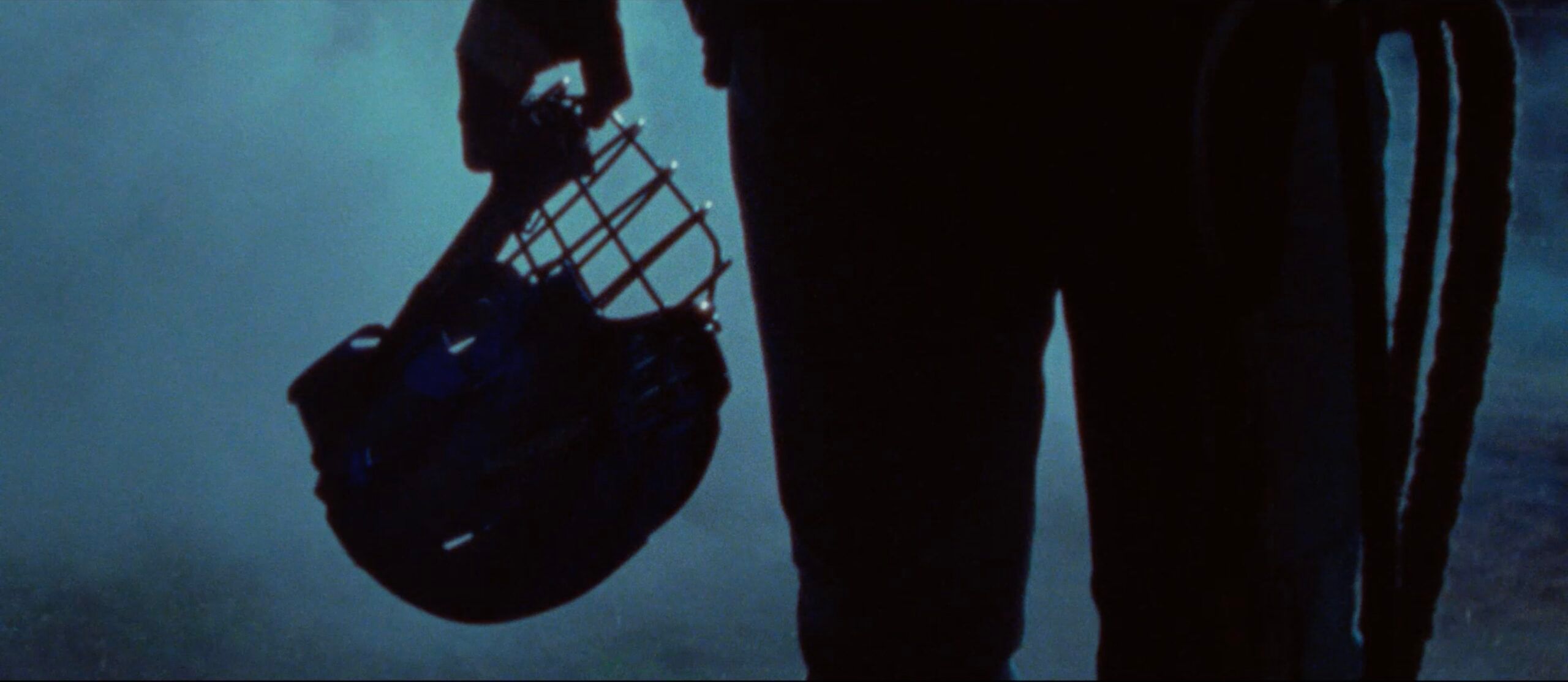 Silhouette of person holding a baseball helmet with protective cage in moody blue light