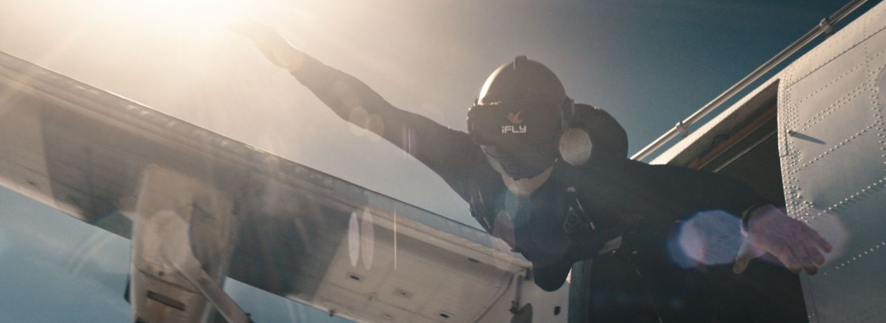 Skydiver exiting aircraft with arms outstretched against bright sun, iFly VR project still.