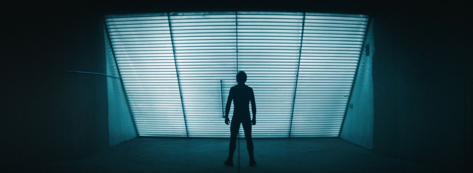 Silhouette of a person standing in front of a large illuminated window with horizontal slats.