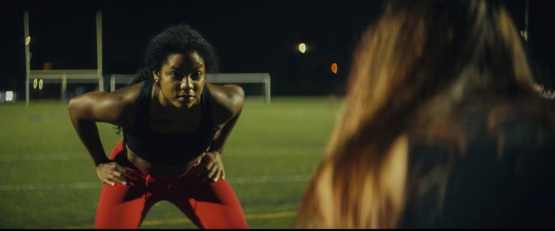 Female athlete in red pants stands on a football field at night facing another player.