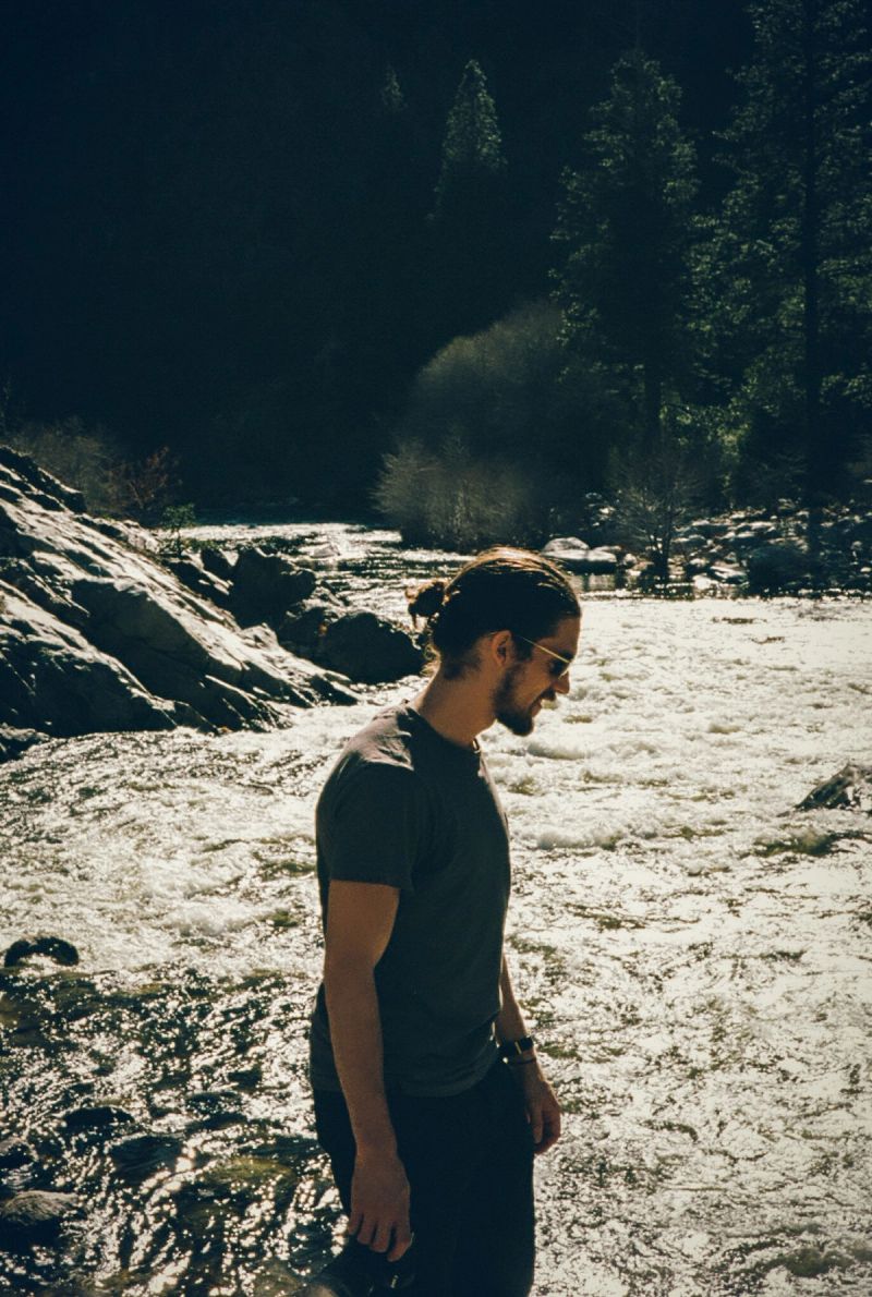 Man in dark t-shirt and sunglasses stands by rushing river with rocky banks and forested hills