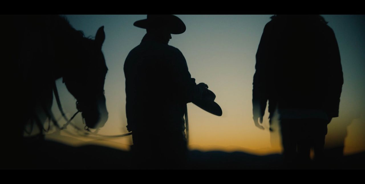 Silhouetted cowboy holding a hat with horse and figure against sunset.
