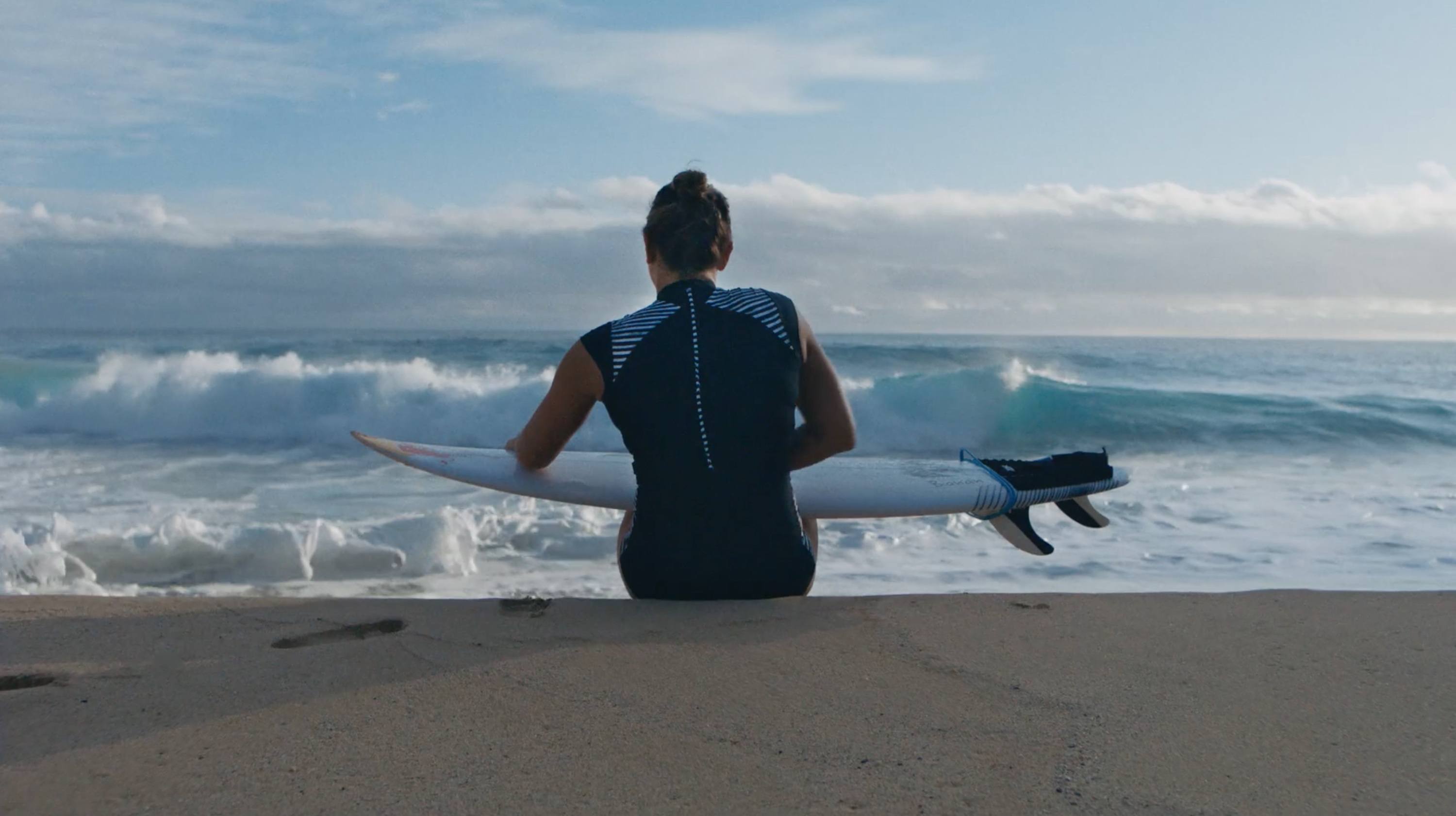 Surfer in wetsuit sits on sand holding surfboard facing ocean waves