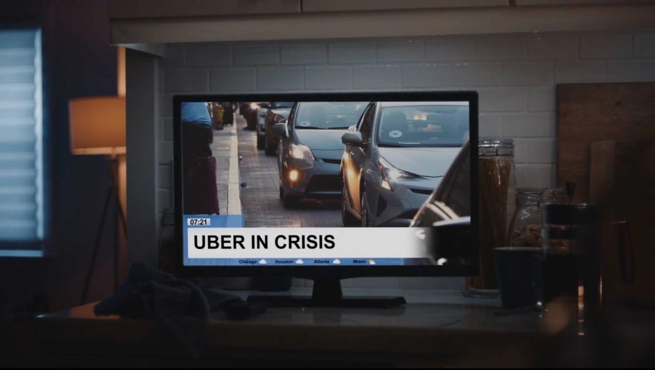 A television screen displays a news report with the headline 'UBER IN CRISIS' over footage of cars.