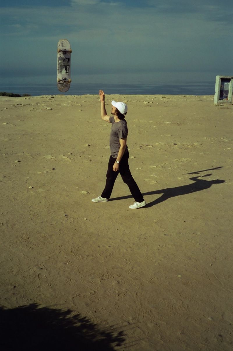 Man in white cap tosses skateboard on sandy beach with ocean backdrop