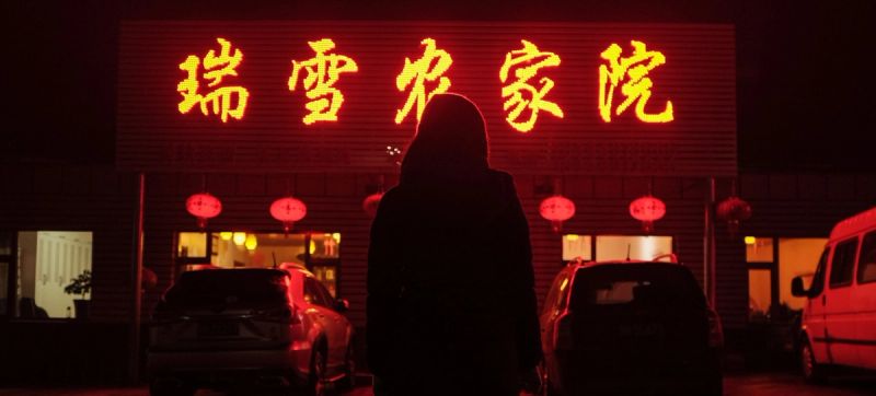 Silhouette of a person standing before a glowing red Chinese sign at night with parked cars.