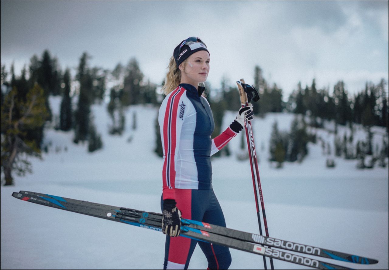 Female skier in red, white and blue suit holding skis and poles on snowy mountain.