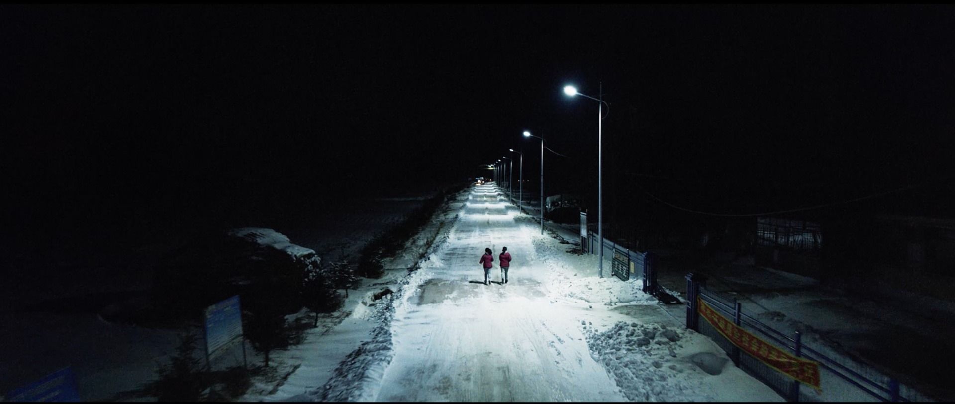 Two figures walk down a snow-covered road at night under streetlights