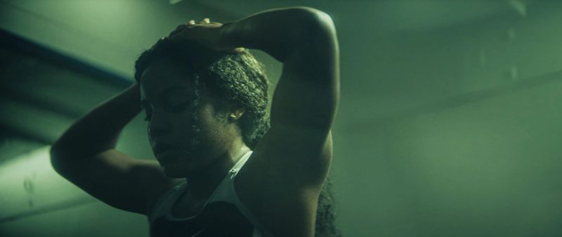 Female athlete stretching with hands behind head in moody green-lit indoor setting.
