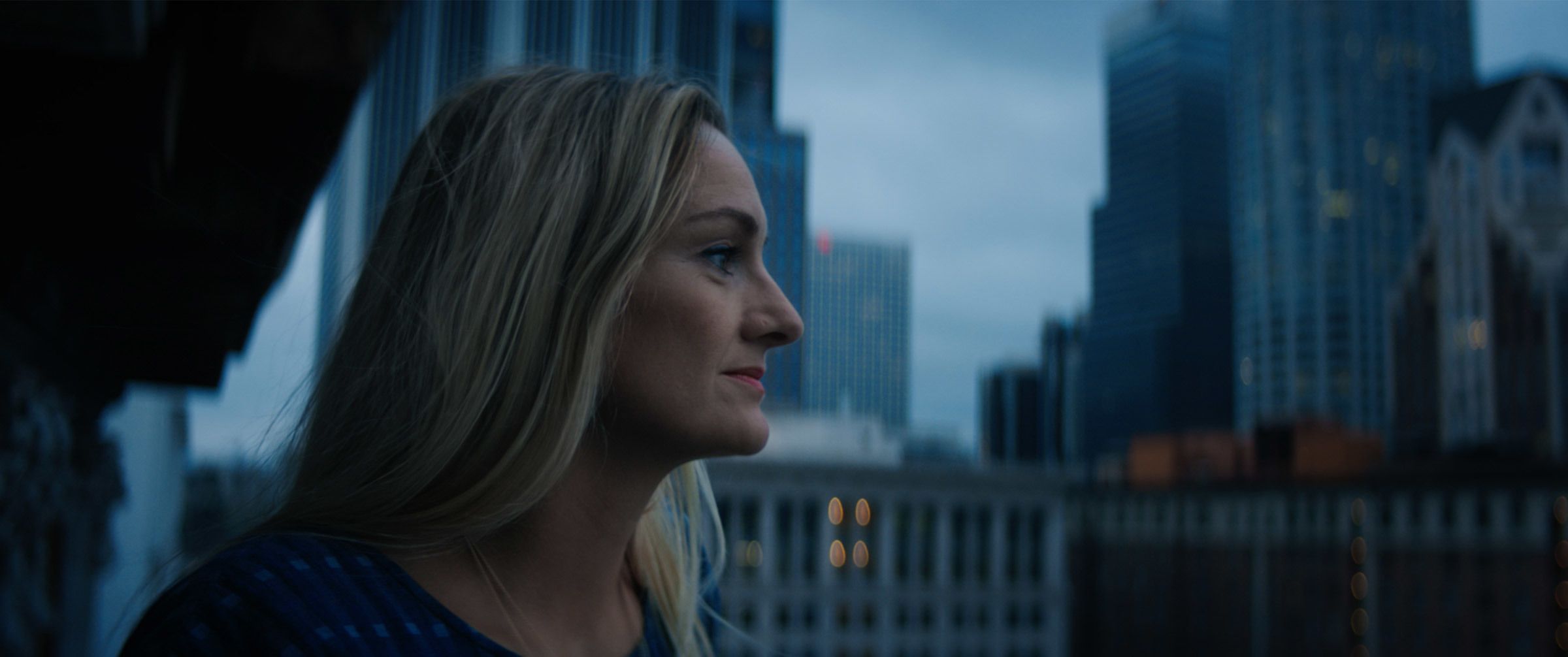 Blonde woman in profile looking out over a blurred city skyline.