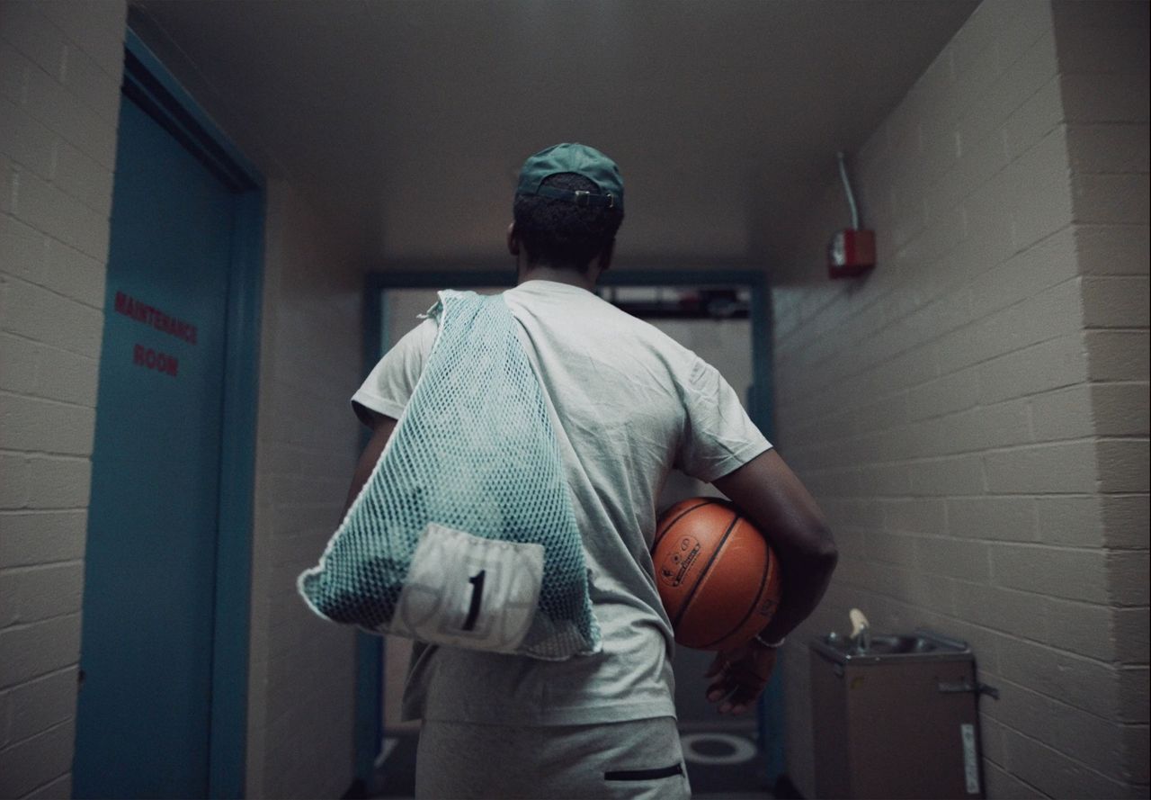 Reggie Jackson walking down a hallway carrying a basketball and mesh laundry bag.