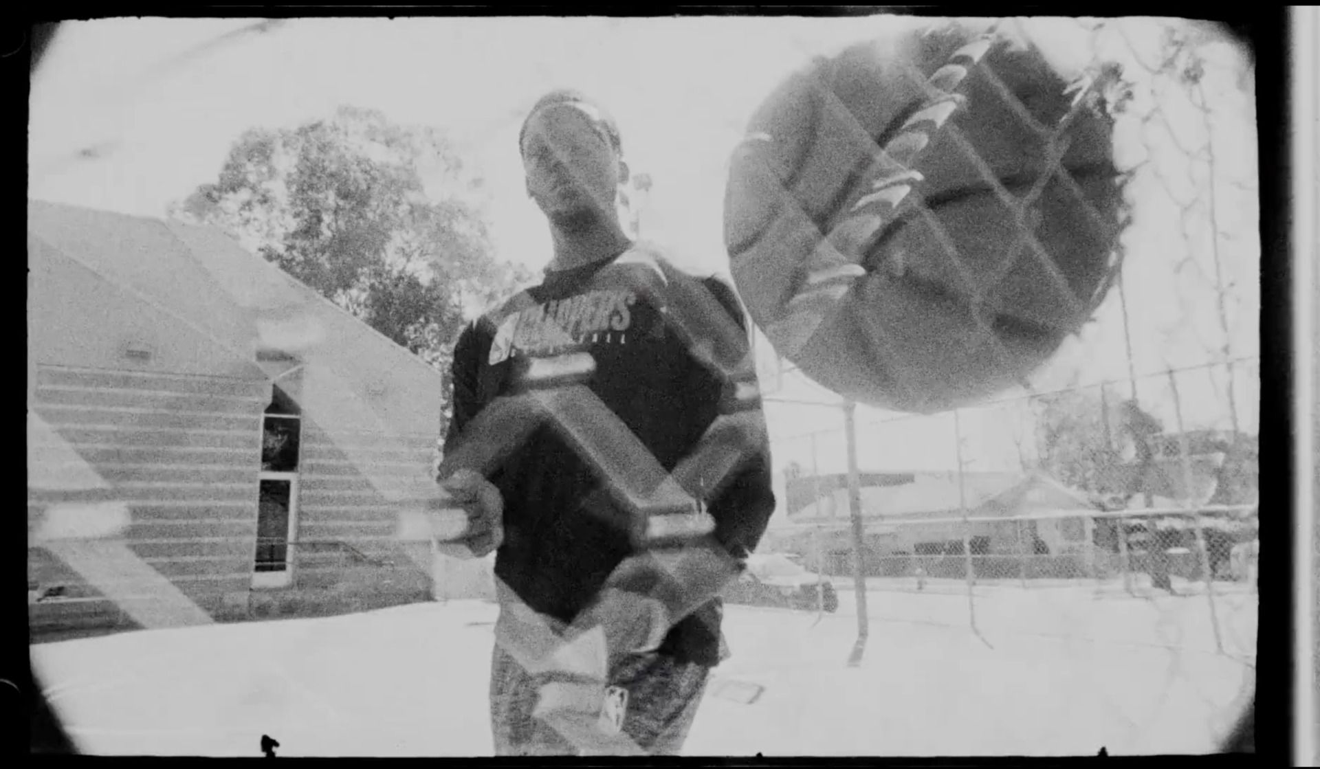 Reggie Jackson holding a basketball outdoors with a chain-link fence overlay, captured in black and white film style.