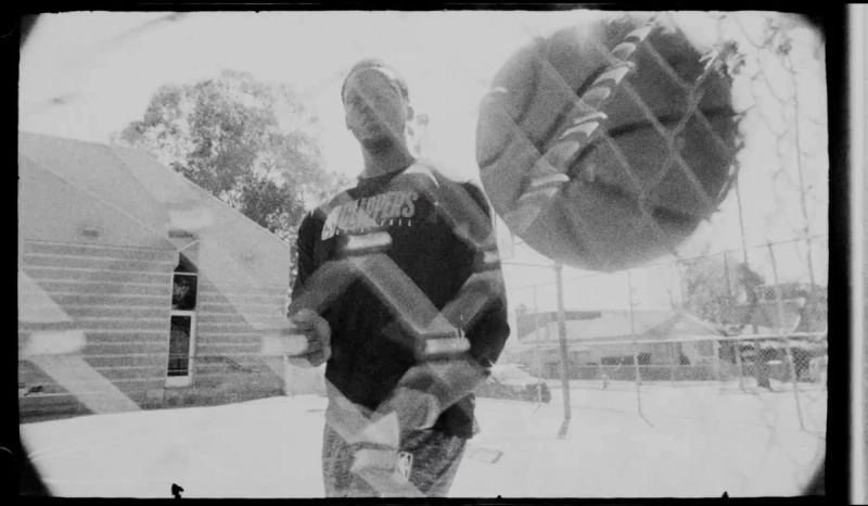 Reggie Jackson holding a basketball outdoors with a chain-link fence overlay, captured in black and white film style.