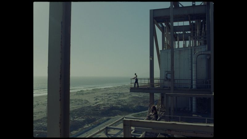 Two figures on an industrial structure overlooking a coastal landscape