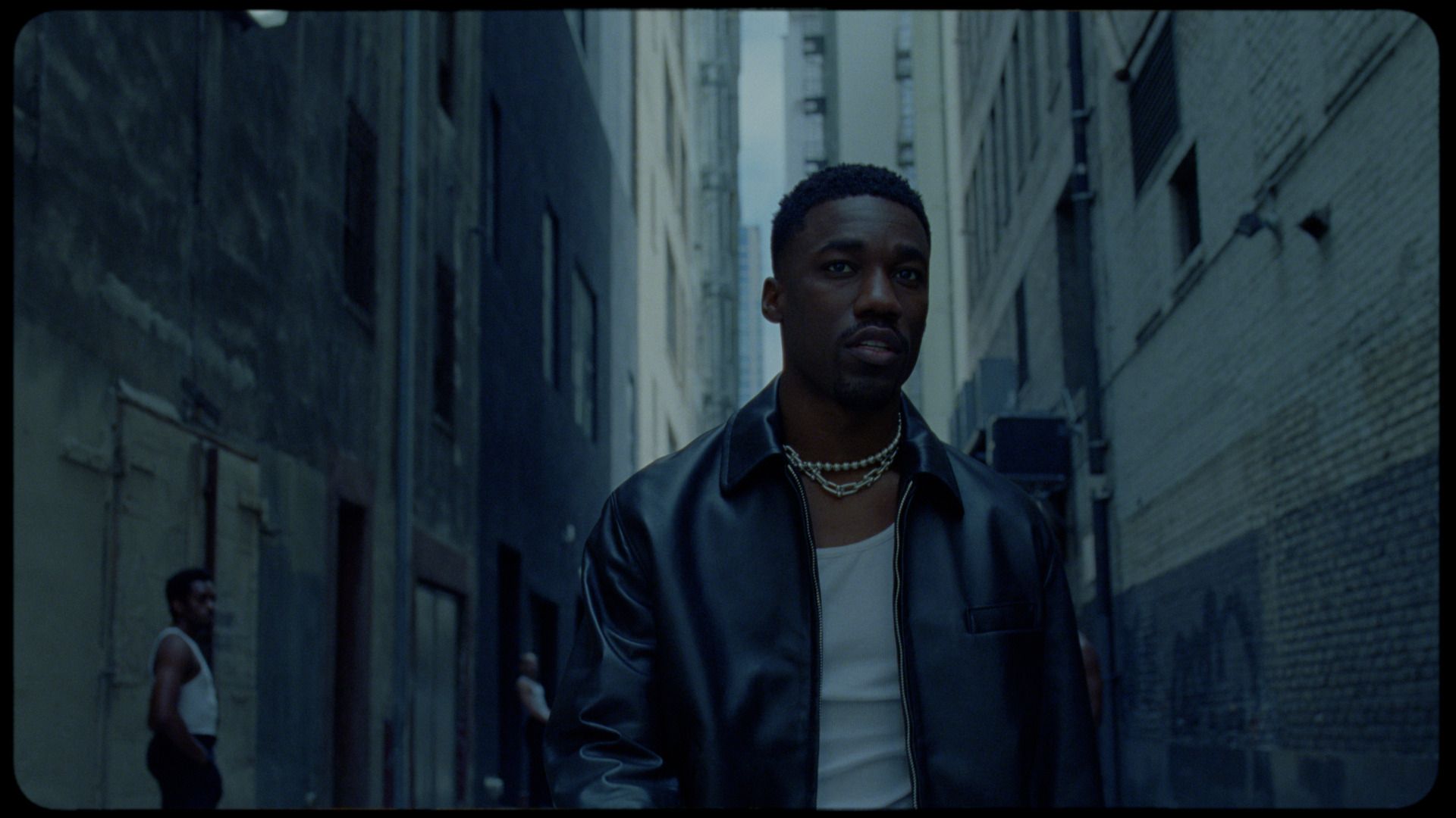 Giveon in a dark leather jacket and layered chains, standing in an urban alleyway.