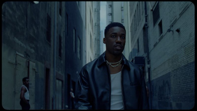 Giveon in a dark leather jacket and layered chains, standing in an urban alleyway.
