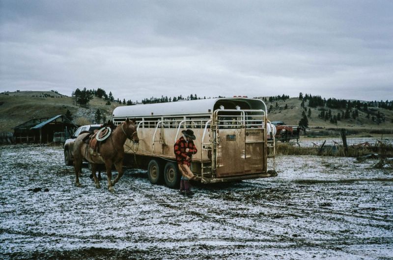 Cowboy stands beside a horse and livestock trailer in a snowy, rural landscape with hills.