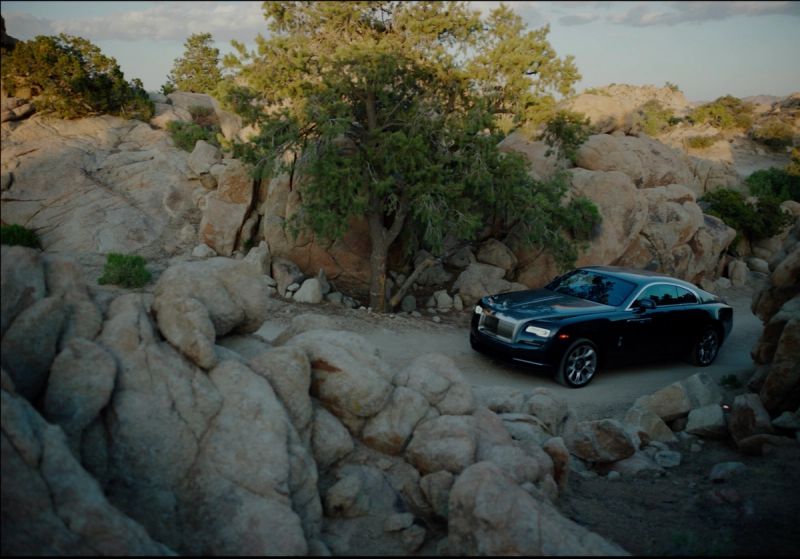 A black Rolls-Royce coupe drives along a winding dirt path through a rocky desert landscape.