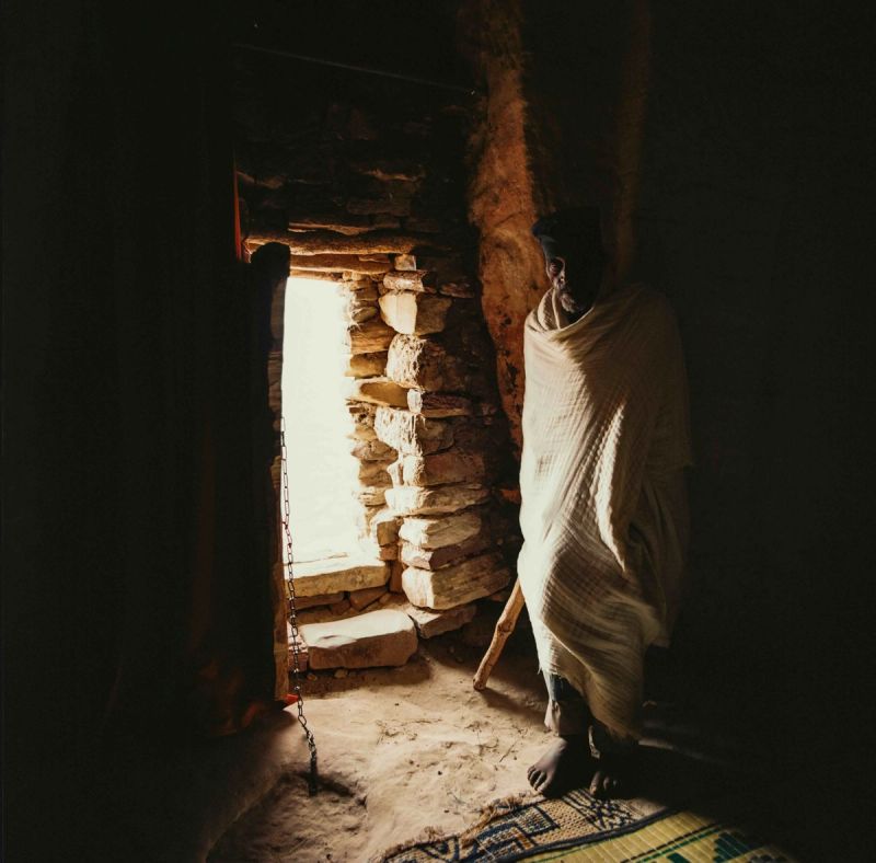Elderly man wrapped in cloth stands in dim light near stone doorway